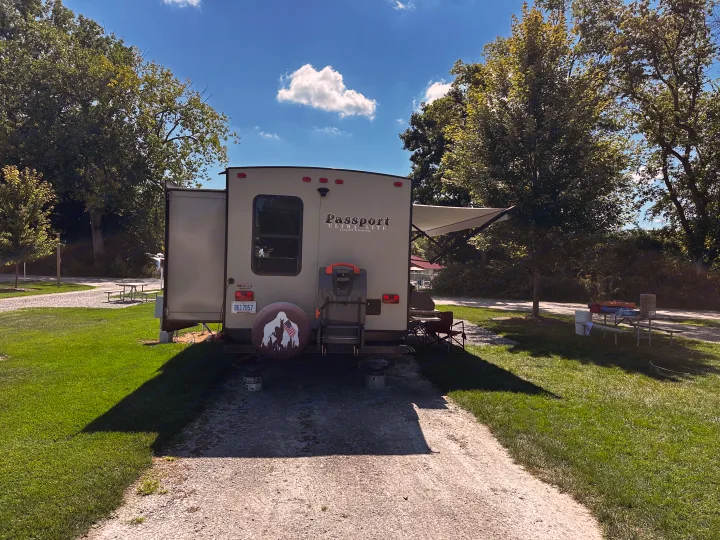An RV rental travel trailer is set up at a sunny campground with its awning extended and a Bigfoot tire cover.. Keystone RV Passport Grand Touring 2017