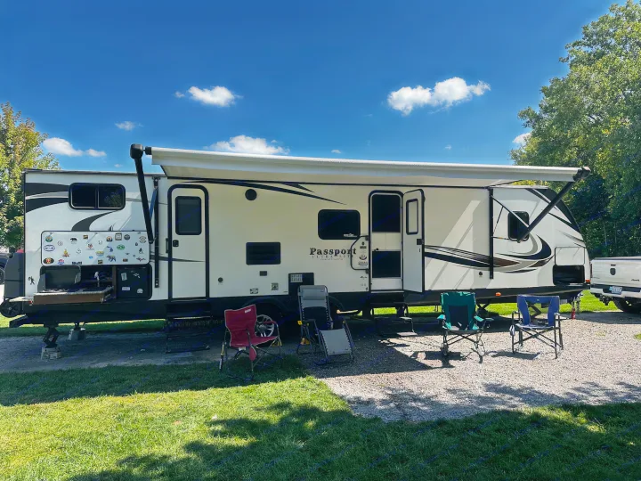 An RV rental campsite with a white travel trailer, extended awning, and multiple camping chairs on gravel and grass. Features an outdoor kitchen.. Keystone RV Passport Grand Touring 2017