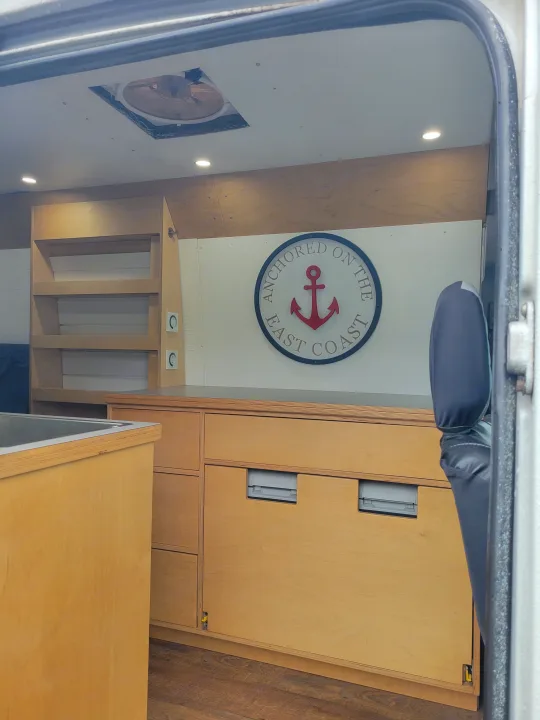 Spacious RV rental van interior featuring wood cabinetry, sink, shelving, and a coastal 'Anchored' sign. Includes ceiling fan.. Mercedes Sprinter 2006