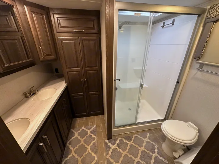 Spacious RV rental bathroom featuring a double vanity with two sinks, dark wood cabinets, a glass shower with a bench, and a toilet.. Keystone RV Alpine 2021
