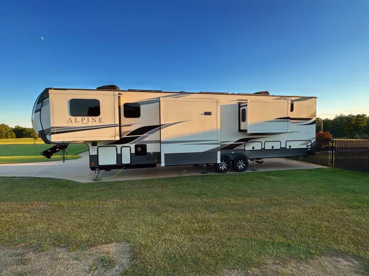 A spacious Alpine fifth-wheel RV rental is perfectly set up on a concrete pad with green grass, under a clear blue sky featuring a crescent moon.. Keystone RV Alpine 2021