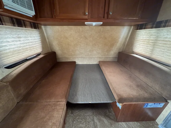 Cozy RV rental interior: dinette with brown upholstered benches, gray table, overhead cabinets, & pleated window blinds.. Jayco Jay Flight Swift SLX 2012