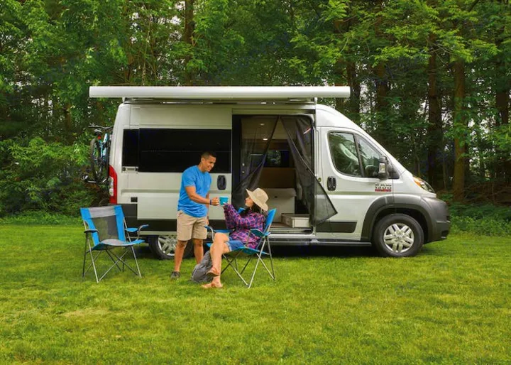 A couple enjoys coffee by their camper van RV rental, parked in a grassy forest setting. Ideal for outdoor adventures.. Thor Rize 2023