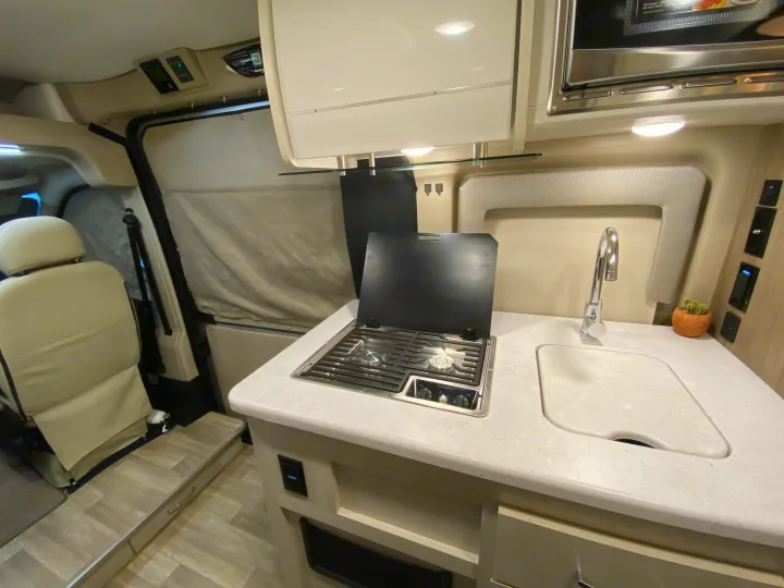 Inside an RV rental, showing a compact kitchen with a two-burner stove, sink, microwave, and light-colored countertops.. Thor Rize 2023