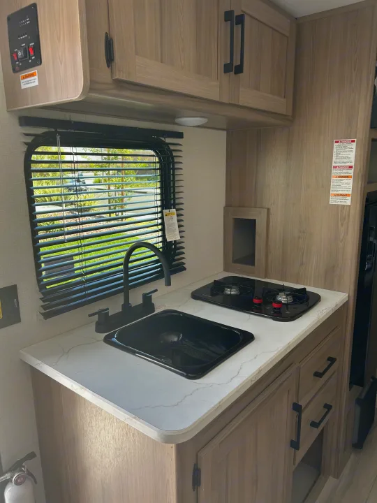 Modern RV rental kitchen with a black sink, two-burner stove, and light wood cabinets, ideal for your next travels.. Coleman Lantern 17B 2025