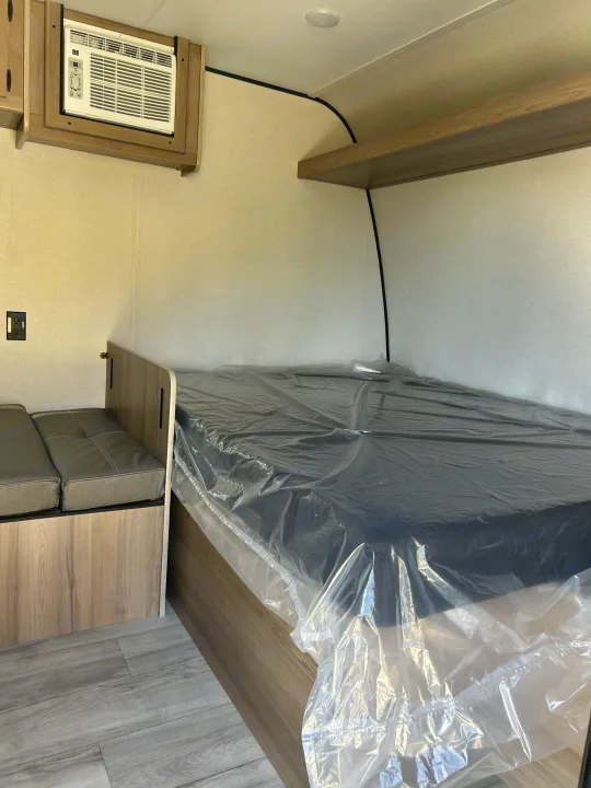 View inside an RV rental showing a plastic-wrapped bed, seating, wall-mounted AC, and wood-look flooring.. Coleman Lantern 17B 2025