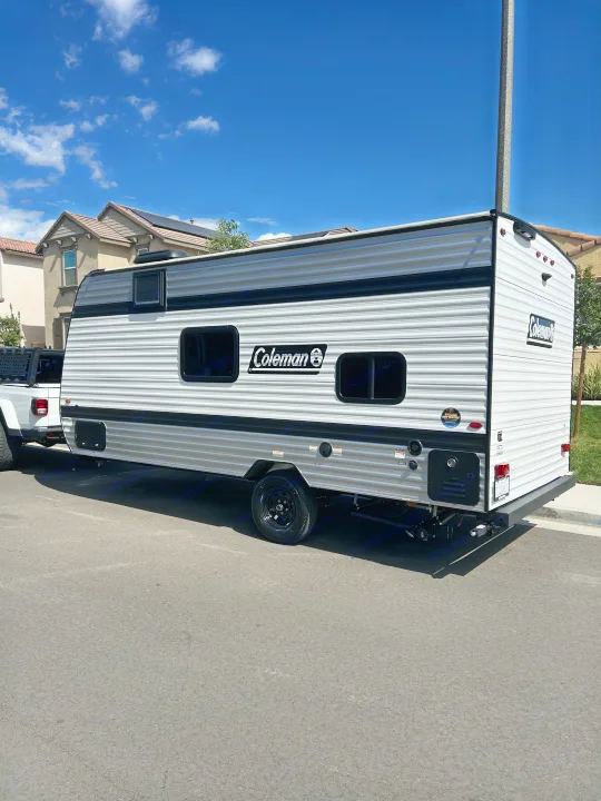A white Coleman travel trailer (RV rental) parked on an asphalt street, towed by a white truck under a sunny blue sky.. Coleman Lantern 17B 2025