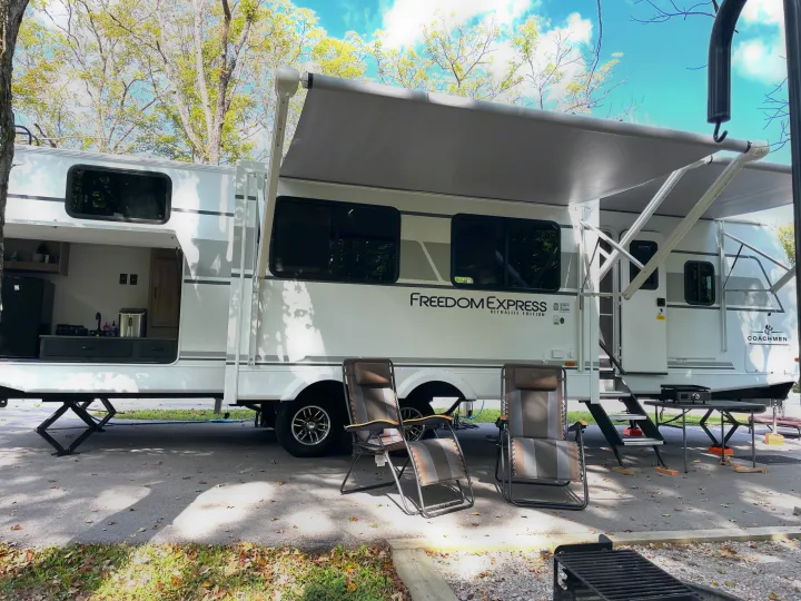 Spacious RV rental at a sunny campsite, complete with an outdoor kitchen, extended awning, and comfortable camping chairs.. Coachmen Freedom Express Ultra Lite 2025