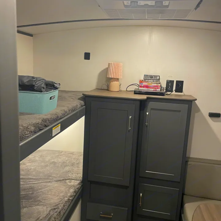 Inside an RV rental: cozy bunk beds with gray blankets, dark cabinets, a lamp, and entertainment setup.. Keystone RV Bullet Ultra Lite 2022