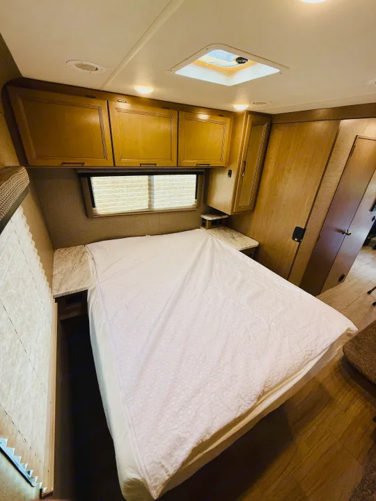 Inside an RV rental, this cozy bedroom features a white bed, wooden cabinets, a window with blinds, and a ceiling skylight.. Thor Coleman 2021