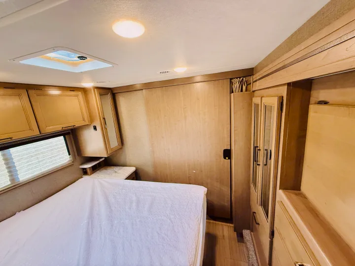 RV rental bedroom showing a white bed, window, light wood cabinets, overhead skylight, and mirrored closet.. Thor Coleman 2021