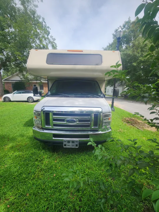 White Ford Class C motorhome, an RV rental, parked on a green lawn with a white convertible and house behind.. Coachmen Leprechaun 220QB 2019