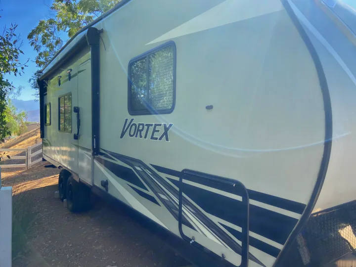 Vortex RV rental parked outside. Light-colored travel trailer with an awning, windows, and graphic decals. Sunny outdoor scene.. Genesis Supreme Rv Genesis Supreme 2022