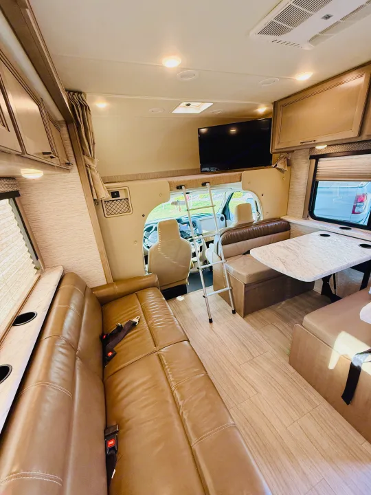 Spacious rv rental interior with a brown leather sofa, dinette, overhead TV, and front cab area visible.. Thor Coleman 2021