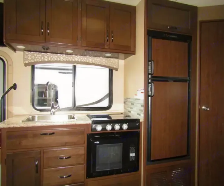 Compact RV rental kitchen with sink, stove, microwave, and fridge, showcasing dark wood cabinetry and a window.. Thor Chateau 2019