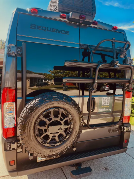 Rear of a dark gray Thor Sequence RV, with spare tire and gear rack, featuring an Outdoorsy sticker for RV rental.. Thor Sequence 2024