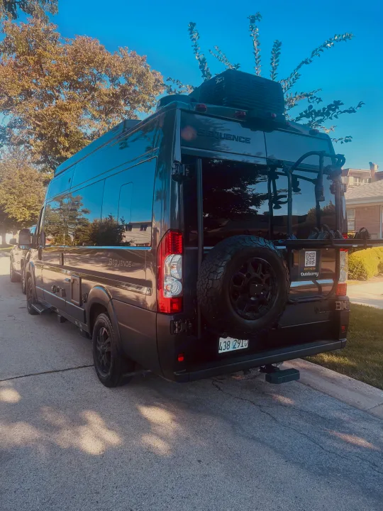 Dark gray Sequence camper van, ready as an RV rental, parked on asphalt with a spare tire and bike rack.. Thor Sequence 2024