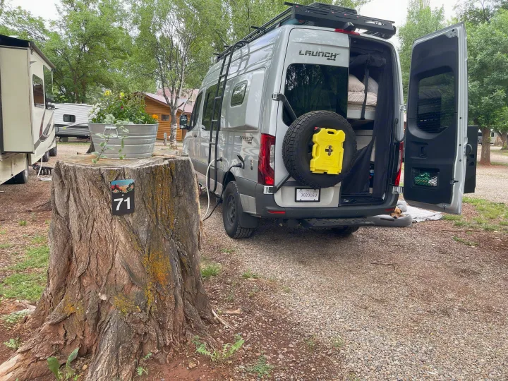 This RV rental camper van is set up at campsite 71, with its rear doors open revealing a cozy interior and a curious dog.. Entegra Coach Launch 2023