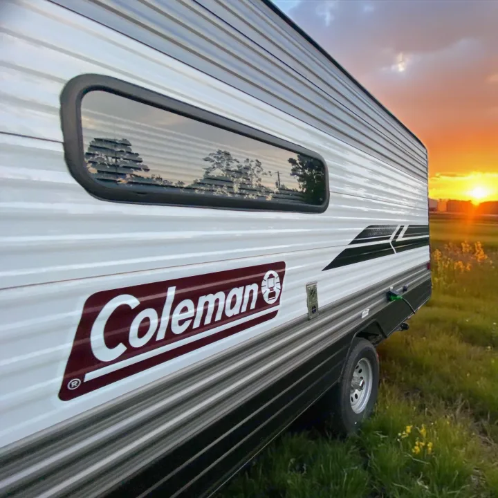 Side view of a white Coleman RV rental, parked in a grassy field with yellow flowers at sunset. Trees reflect in the window.. Dutchmen Coleman Lantern LT 2022