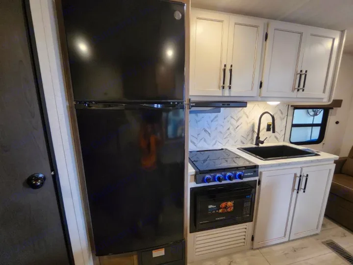Modern RV rental kitchen with a tall black fridge, stovetop, microwave, black sink, and white cabinets/backsplash.. Keystone Springdale Classic 2025
