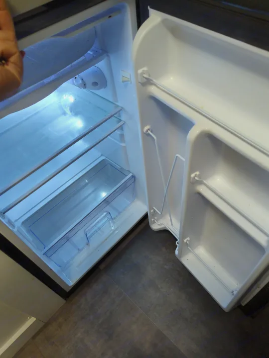 Open, empty compact refrigerator in an RV rental, showing glass shelves and door compartments.. Travel Lite Rove Lite 2023