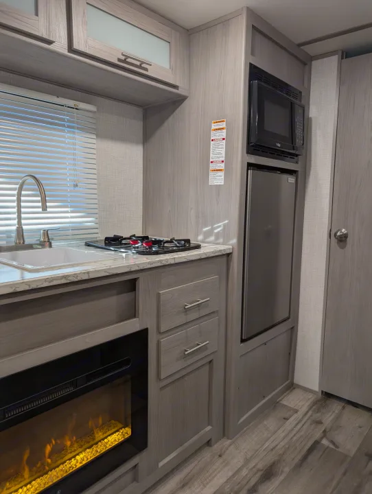 Modern RV rental kitchen featuring a sink, stove, microwave, fridge, and built-in electric fireplace with light wood cabinetry.. Dutchmen Coleman Lantern LT 2023