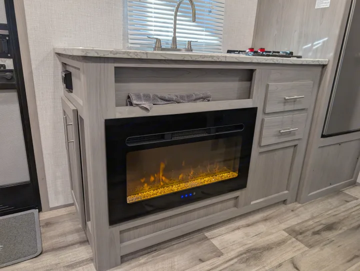 This modern RV rental features a kitchenette with a sink, stovetop, and an electric fireplace built into light wood cabinetry.. Dutchmen Coleman Lantern LT 2023