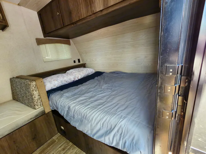 Cozy RV rental bedroom featuring a made bed with pillows, a window with blinds, and wood-look overhead cabinets.. Forest River Viking 2020
