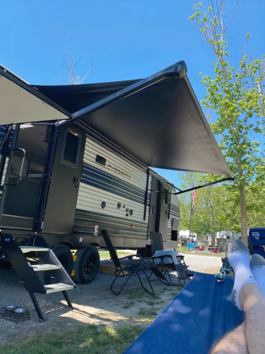 Relaxing with feet up under the extended awning of a dark gray RV rental, parked at a sunny campsite with pet bowls nearby.. Forest River Cherokee 2021