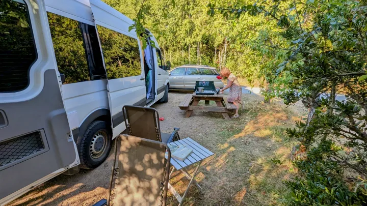 Sunny outdoor cooking scene with an RV rental camper van. A woman cooks on a picnic table; lounge chairs are set up nearby.. Mercedes Sprinter 2008
