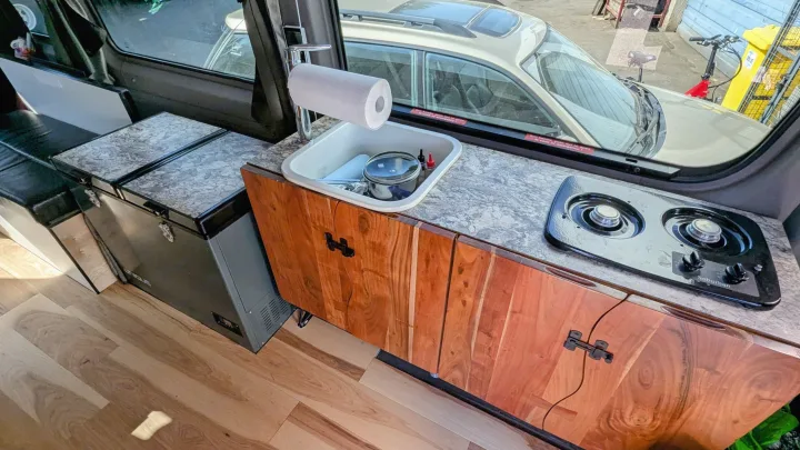 Inside an RV rental, showing a compact kitchen with a sink, two-burner stove, refrigerator, and wooden cabinets. Outside, another vehicle and bike are visible.. Mercedes Sprinter 2008