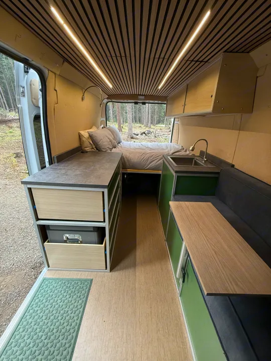Inside a modern rv rental, featuring a comfortable rear bed, full kitchen, and flexible living space with wood accents.. Dodge B Van 2023