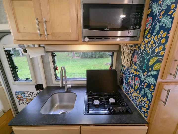 RV rental kitchen: wood cabinets, microwave, sink, stove, plus patterned wall decor & outdoor view.. Xtreme Outdoors Little Guy Max 2021