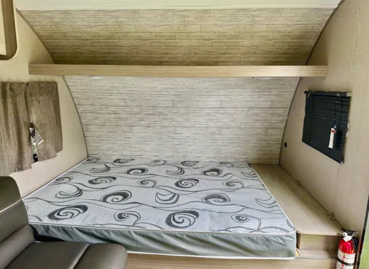 Cozy RV rental interior with a patterned mattress, wood-look walls, and compact sleeping area. Features a window with blinds.. Coachmen Catalina 2023
