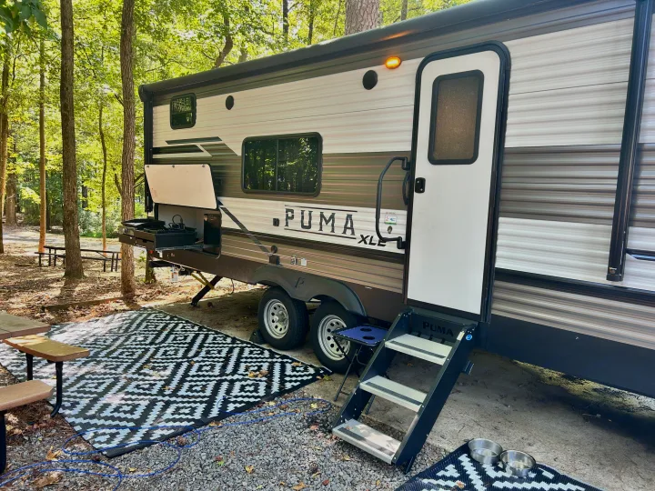 PUMA XLE travel trailer setup at a wooded RV rental campsite with outdoor kitchen, patterned rug, and steps. Pet bowls nearby.. Forest River Palomino Puma XLE 2023