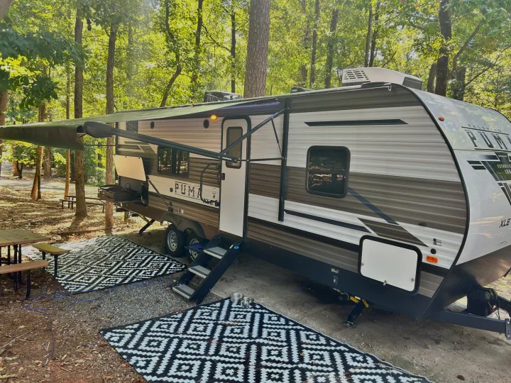This inviting RV rental is comfortably set up at a wooded campsite, featuring an extended awning, outdoor kitchen, and patterned ground mats.. Forest River Palomino Puma XLE 2023