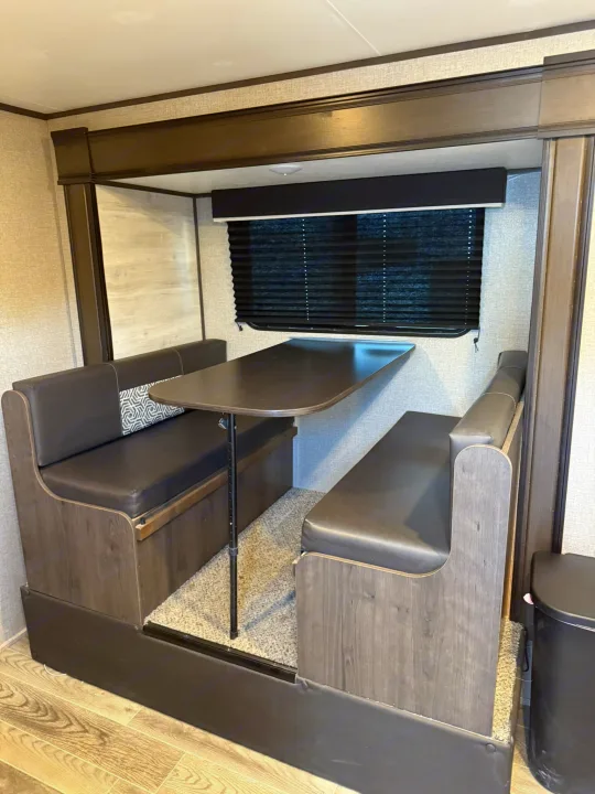An RV rental dinette with dark upholstered booth seating, a central table, and pleated window blinds, offering a cozy interior.. Jayco Jay Flight SLX 2021