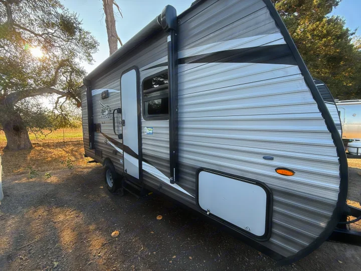 RV rental: Modern Jayco travel trailer with awning, parked on gravel next to a sunlit tree for your trip.. Jayco Jay Flight SLX 2021
