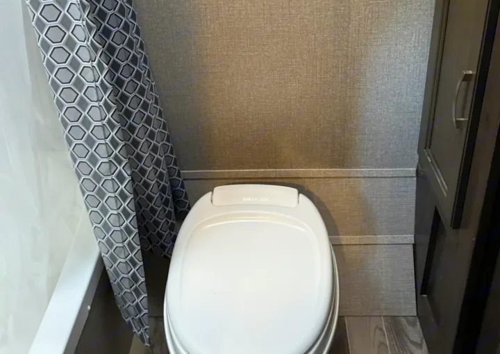Inside an RV rental, showing a compact bathroom with a white toilet, geometric grey shower curtain, and dark wood cabinetry.. Jayco Jay Flight SLX 2021