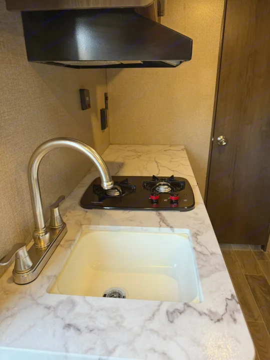 RV rental kitchen: stainless steel faucet, white sink, 2-burner Suburban stove on marble-look counter. Brown walls & door.. Jayco Jay Flight SLX 2021