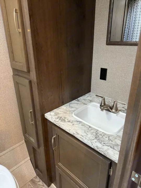 Compact RV rental bathroom with a white sink, marble-look countertop, and dark wood cabinets. A mirror reflects a curtain.. Jayco Jay Flight SLX 2021