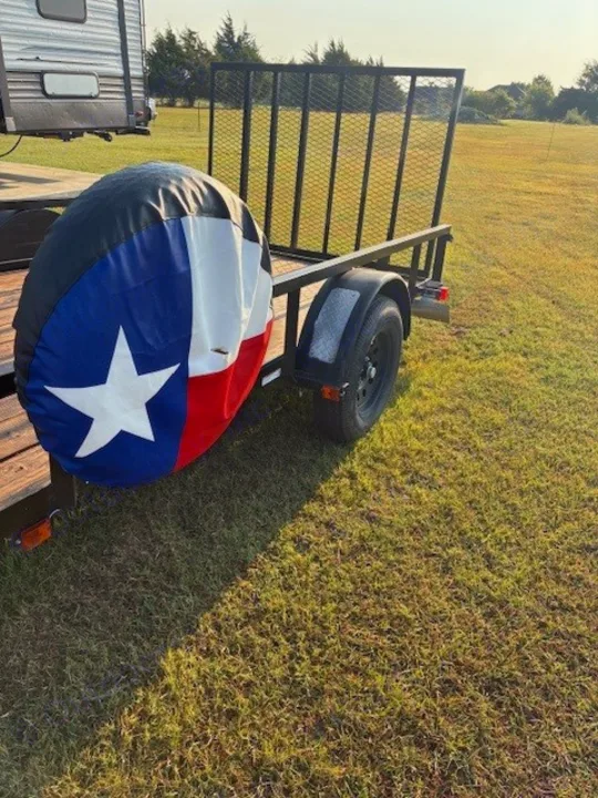 Black utility trailer with a Texas flag spare tire cover; an RV is parked nearby in a grassy field. Ideal for your RV rental trips!. Carry On Trailers 6X12GWPTLED 2022