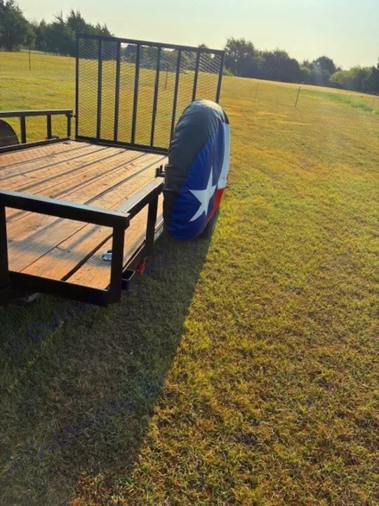 A Texas flag spare tire on a utility trailer parked in a sunny field, ready for your RV rental adventures.. Carry On Trailers 6X12GWPTLED 2022