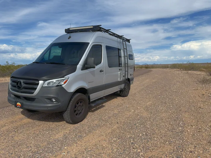 Silver Mercedes Sprinter RV rental, equipped with a roof rack and ladder, parked on a gravel desert road.. Entegra Coach Launch 2023