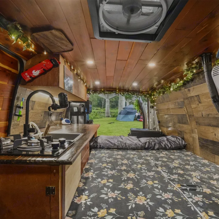 Cozy wood-paneled RV rental interior with kitchen, floral bed, and open back revealing a grassy campsite with tents.. Ford Econoline 150 2007