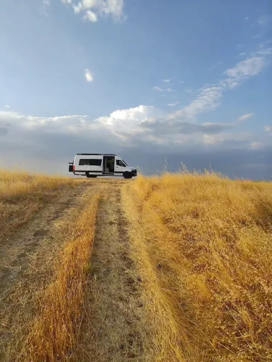 RV rental adventure! White camper van on a golden hill track under a partly cloudy blue sky, perfect for outdoor travel.. Custom Crafted Vans Family Van 2019