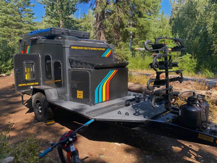 A black Bearlander 20 overland camper with bikes, ideal for shredding, parked in a sunny forest. Consider this RV rental type!. ITWO custom Pixtech Bearlander 20 2024