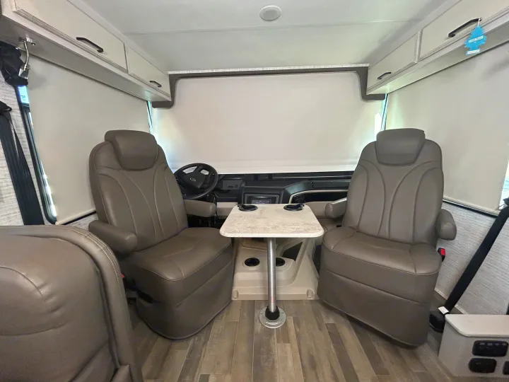 Inside an rv rental, showing the driver's and passenger's seats, a small table, and overhead storage. Window shades are down.. Jayco Alante 2024