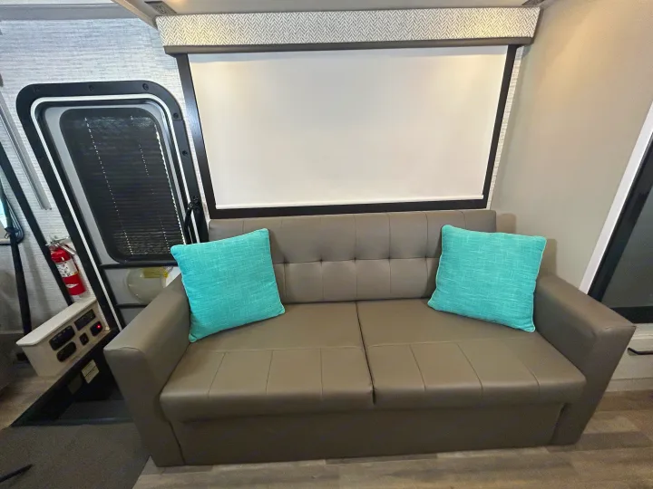 Cozy RV rental interior featuring a gray sofa with teal pillows, a large white screen, and a door with a fire extinguisher.. Jayco Alante 2024