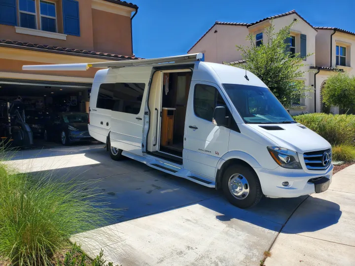 A white Mercedes-Benz Sprinter van RV rental, with an extended awning and open side door revealing its interior, is parked on a sunny driveway.. Coachmen Galleria 2019